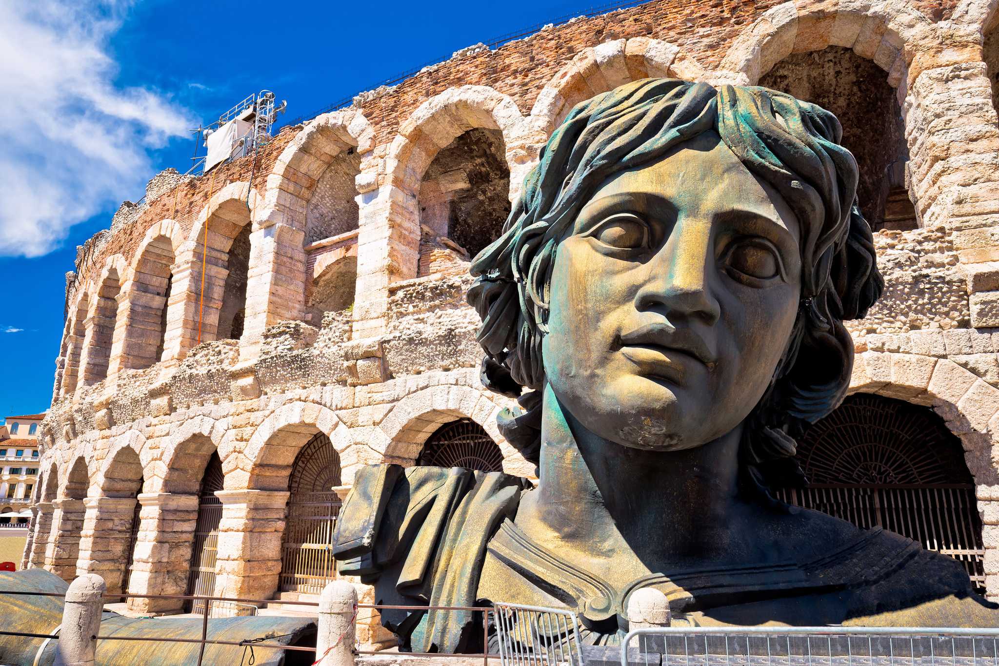 A bronze bust sculpture in front of the arched stone facade of the Verona Arena under a blue sky.