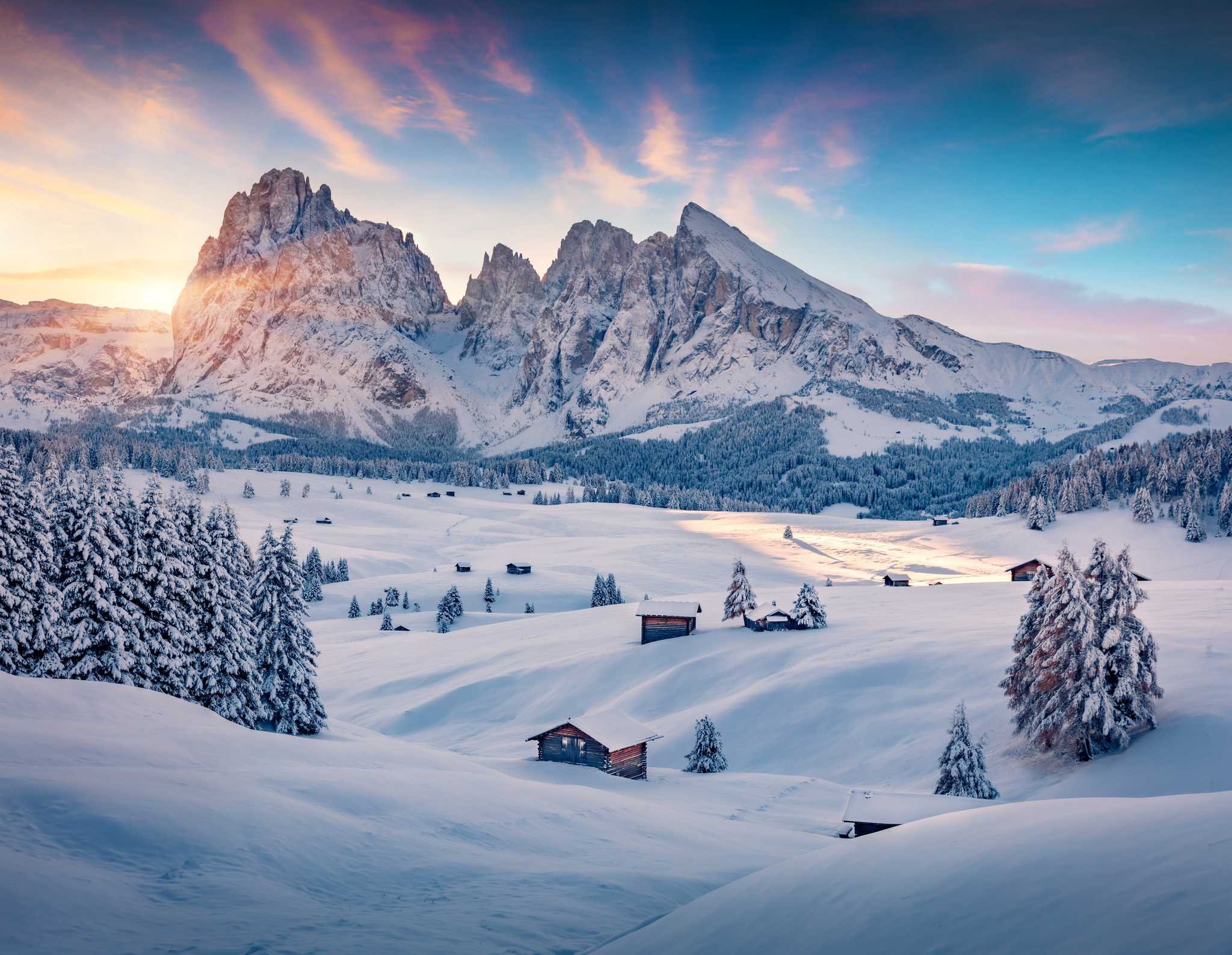 Snow-covered meadow with wooden cabins and the Dolomite peaks at sunrise in winter.