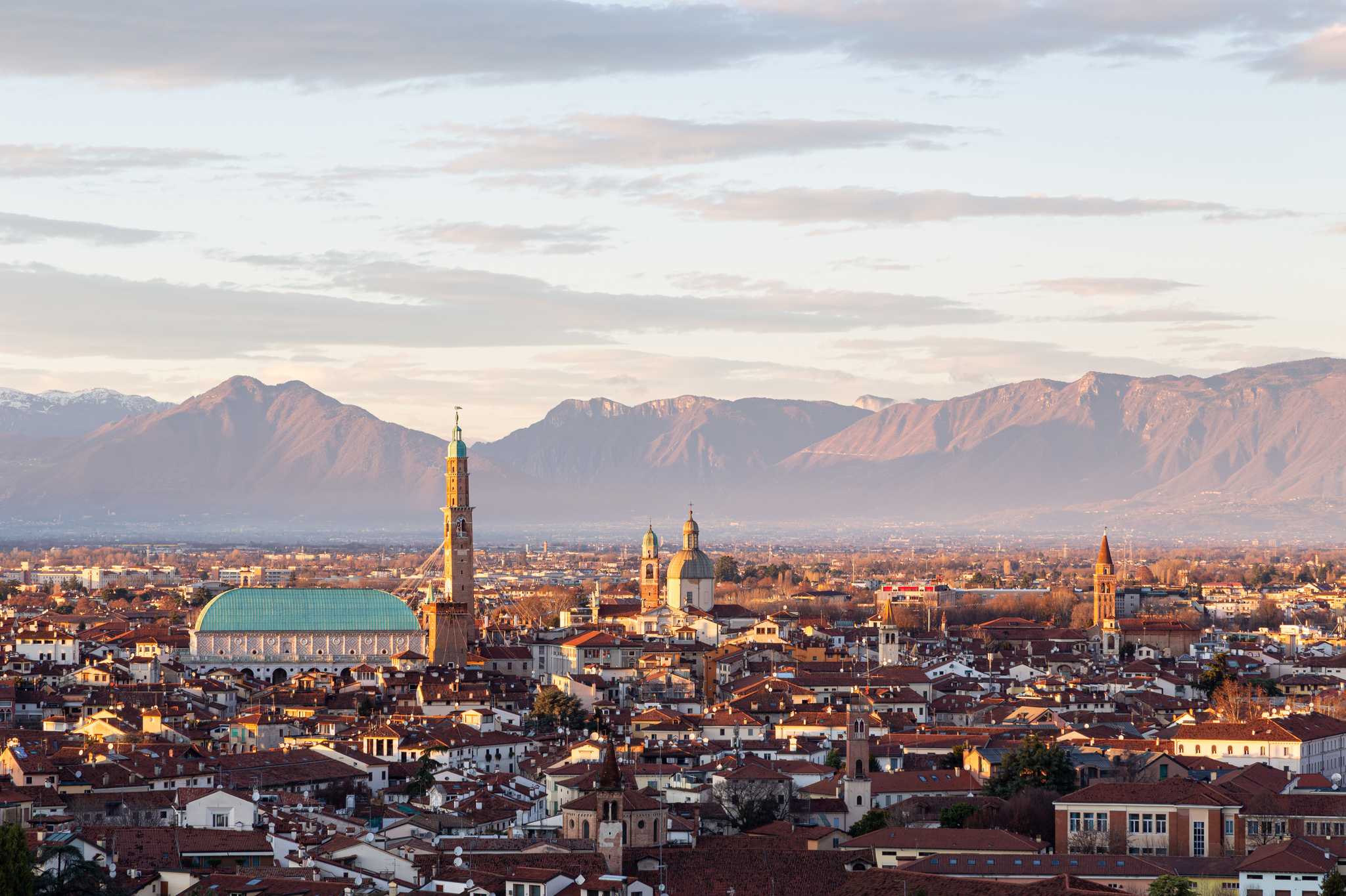 The Vicenza skyline at sunset with the Basilica Palladiana's green roof and tower against a mountain backdrop.
