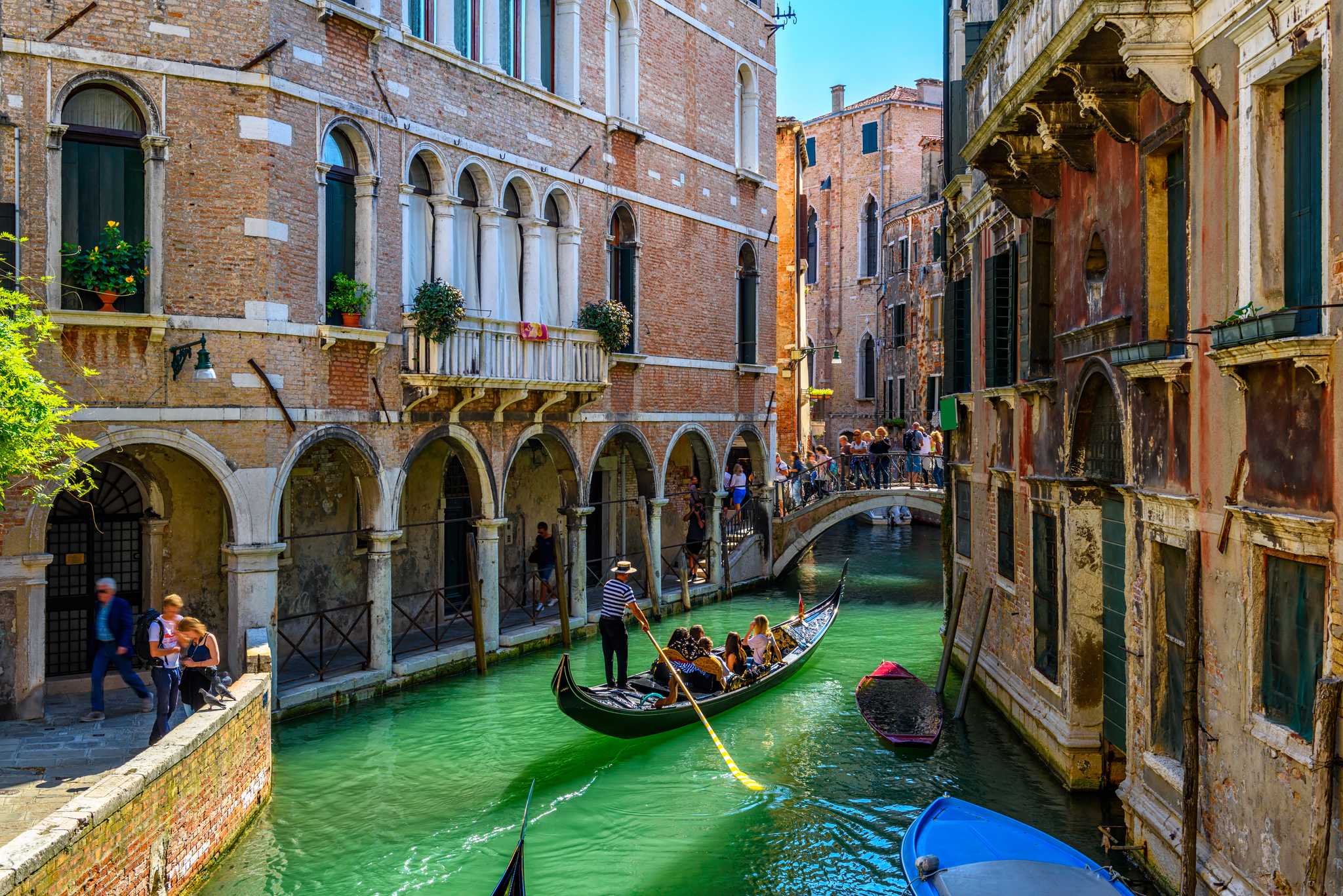 The tight canals of Venice, with a gondola approaching a low bridge.