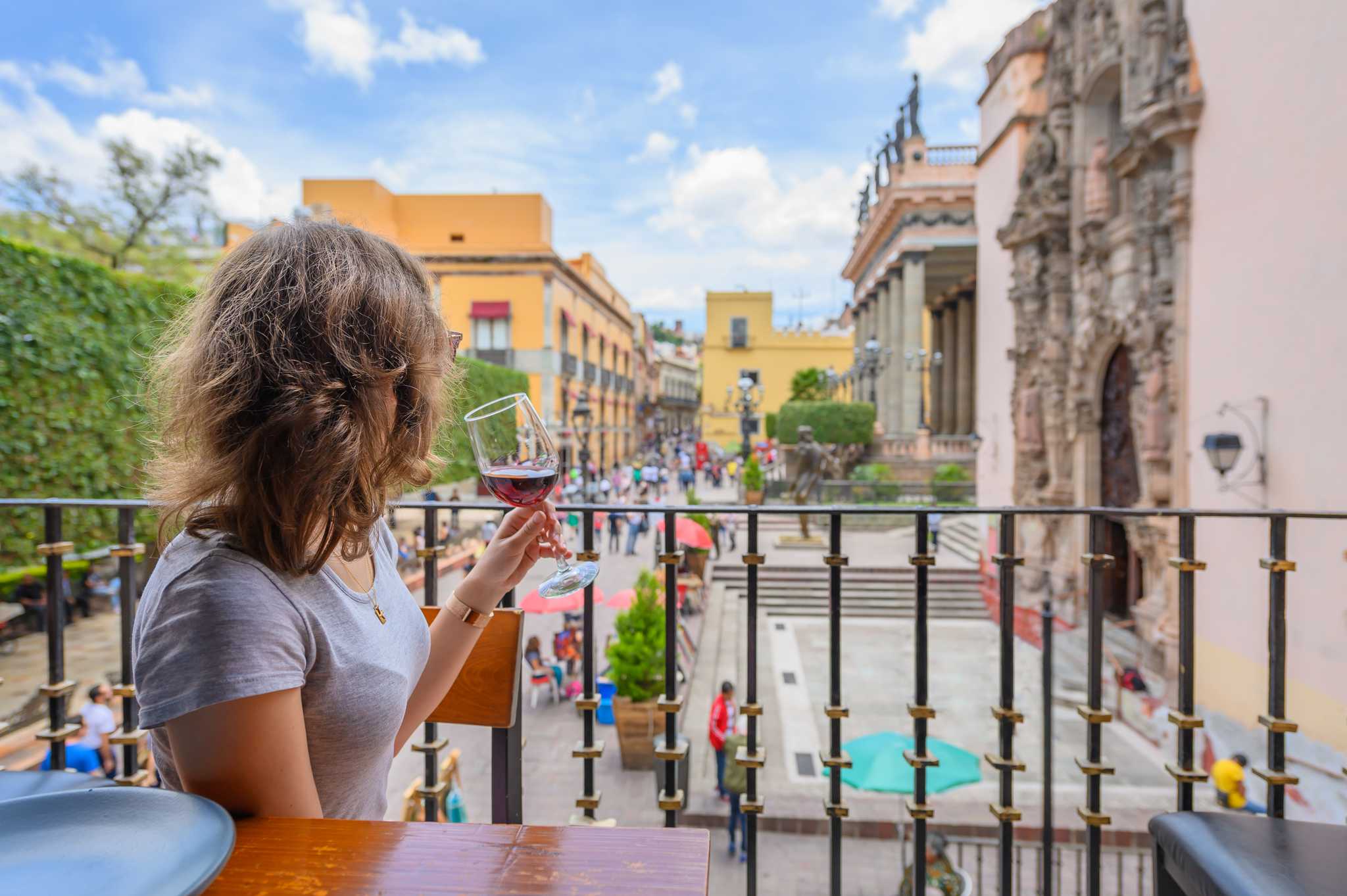 A woman holds a wine glass as she looks over a busy plaza in Mexico on a sunny day.