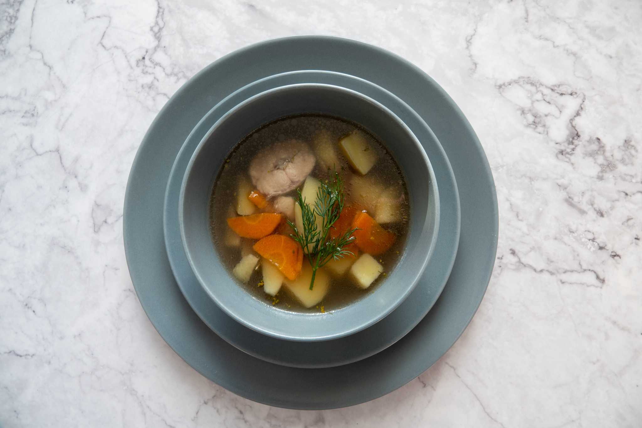 A blue bowl of clear broth with meat, potatoes, carrots, and fresh dill on a marble countertop.